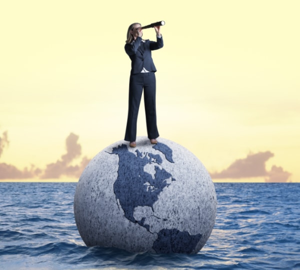 A businesswoman peers through a spyglass while standing on a globe that is adrift in the ocean.